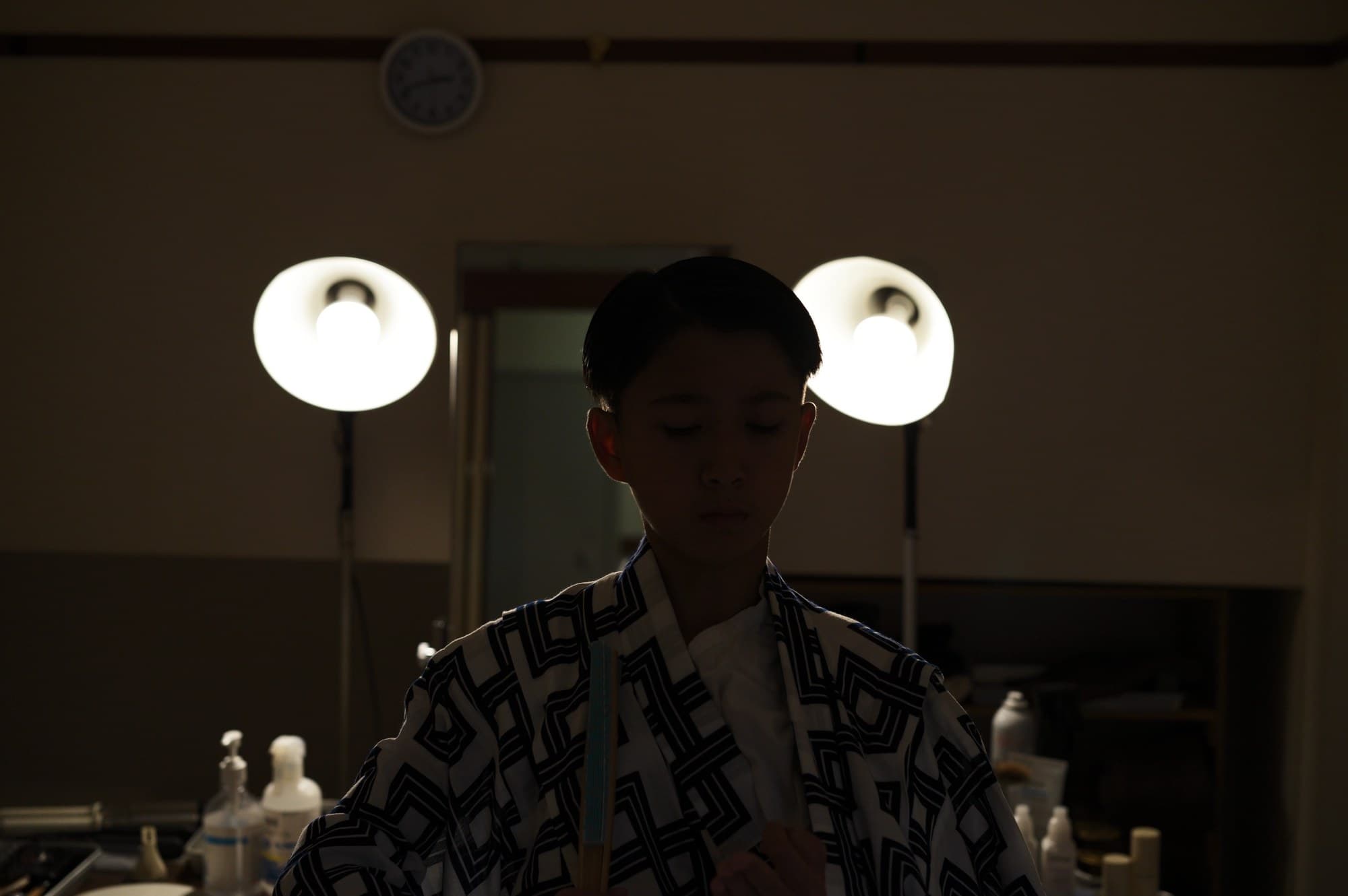 楽屋の丸鏡照明の前に立つ、柄物の着物姿の人物